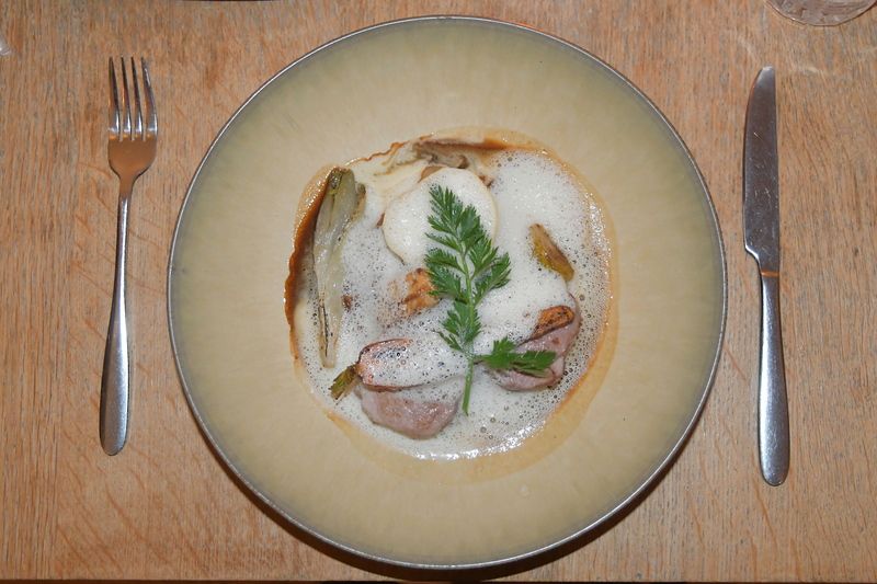 Een bord met vlees, groenten, schuimige saus en een groen takje, met mes en vork ernaast.