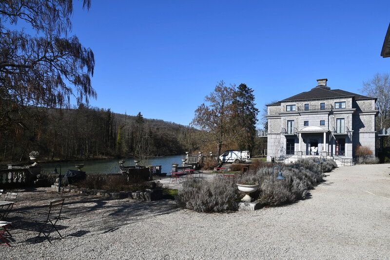 Een statig huis met een grindterras, struiken en tuinstoelen naast een rivier.