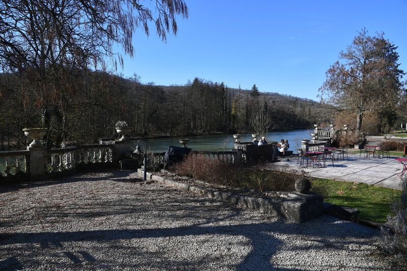 Een terras met grind, tuinbeelden en tafeltjes langs een rivier, met beboste heuvels op de achtergrond.