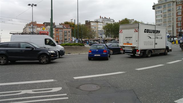 Een kleine blauwe personenwagen wurmt zich nog gauw tussen twee aanstormende auto's en een vrachtwagen op het Meiserplein.