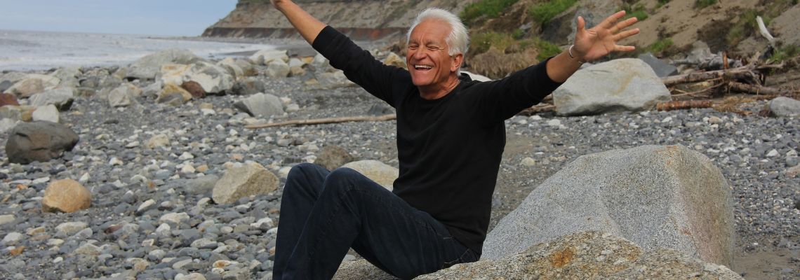 Een lachende oudere man zit op een rots aan het water met zijn armen wijd open.