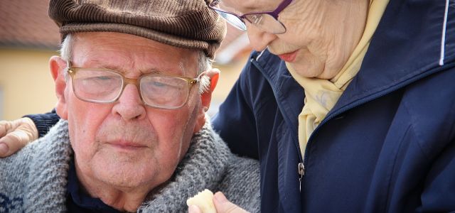 Oudere man met pet en bril naast een vrouw die naar hem toe buigt.