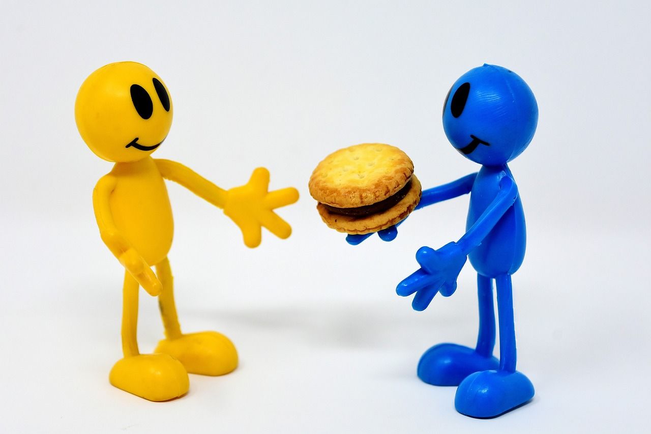 Een blauw speelfiguurtje biedt een koekje aan een geel figuurtje aan.