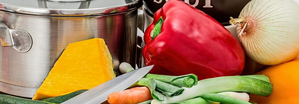 Paprika, ui, pompoen, wortel en lente-ui naast een kookpot en een mes.