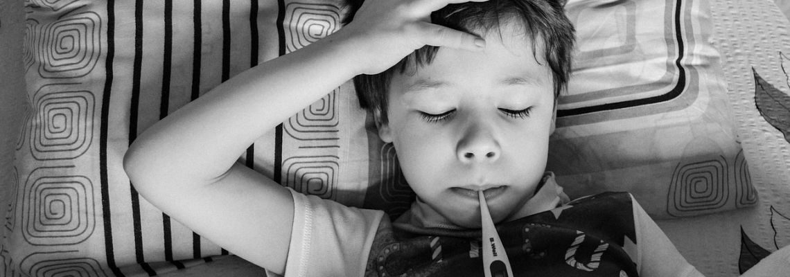 Een kind ligt in bed met een thermometer in de mond en een hand op het voorhoofd.
