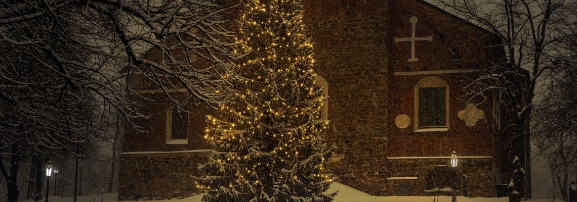 Een grote kerstboom met lichtjes staat in de sneeuw voor een kerk.
