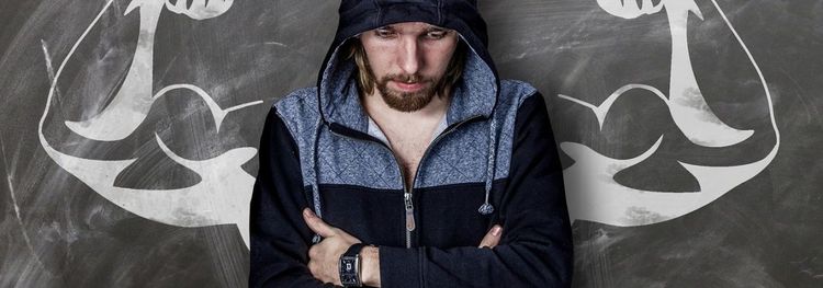 Een man met een kap op kruist zijn armen voor een bord met getekende spierballen.