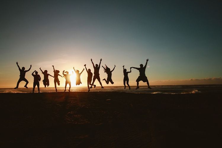 Een groep mensen springt met de armen omhoog op het strand bij zonsondergang.