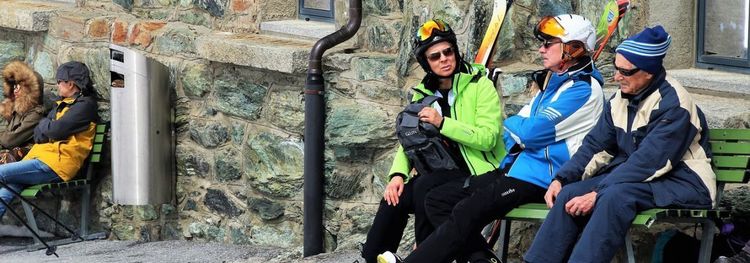 Drie oudere wintersporters zitten met hun ski-uitrusting op een bank tegen een stenen muur.