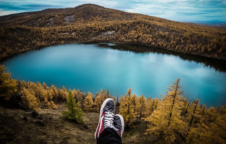 Benen met sportschoenen boven een blauw meer, omringd door heuvels en bomen in herfstkleuren.