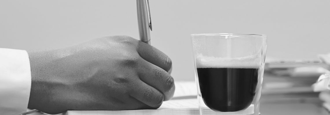 Een hand schrijft met een pen op papier, naast een glas koffie.