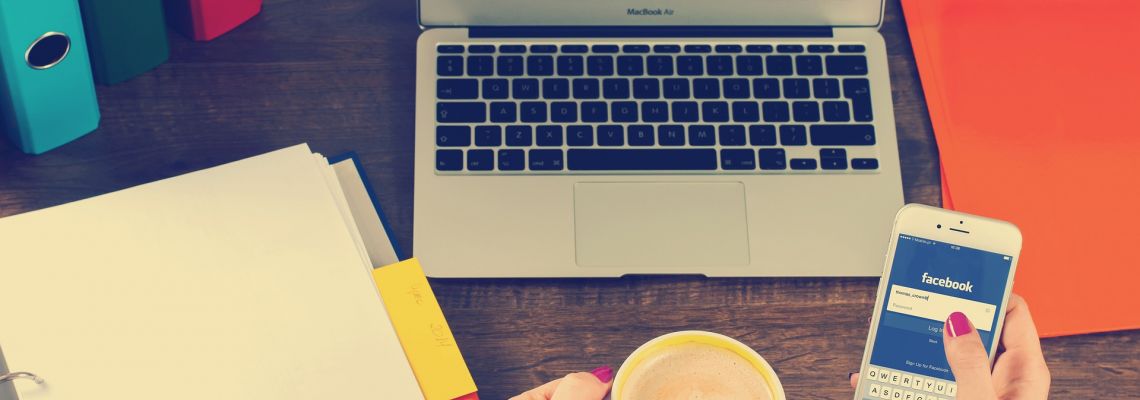Een laptop, papieren en een kopje op een bureau, met een smartphone waarop Facebook openstaat.