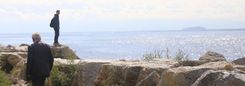 Twee mensen staan op rotsen aan zee, één op de voorgrond en één dichter bij het water.