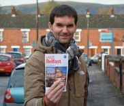 Een man met een sjaal houdt een toeristische brochure over Belfast vast in een straat met bakstenen huizen.