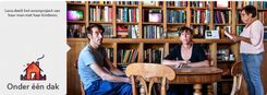 Twee mensen zitten aan een ronde tafel, een derde staat met een boek bij een volle boekenkast.