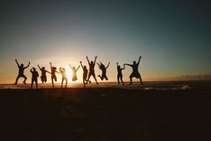 Een groep mensen springt met de armen omhoog op het strand bij zonsondergang.