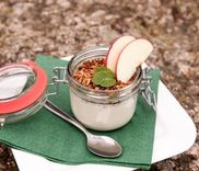 Glazen pot met yoghurt, granola, schijfjes appel en een blaadje munt, met een lepel ernaast.