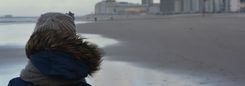 Een persoon met een wollen muts en een warme jas kijkt uit over het strand en de zee.