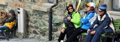 Drie oudere wintersporters zitten met hun ski-uitrusting op een bank tegen een stenen muur.