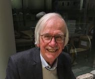 Een oudere man met een bril en een snor glimlacht naar de camera.