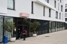 De ingang van een modern hotel met het opschrift YUST, twee mensen en bomen in potten.