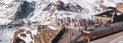 Een rode trein bij een bergstation, met reizigers en besneeuwde bergtoppen.