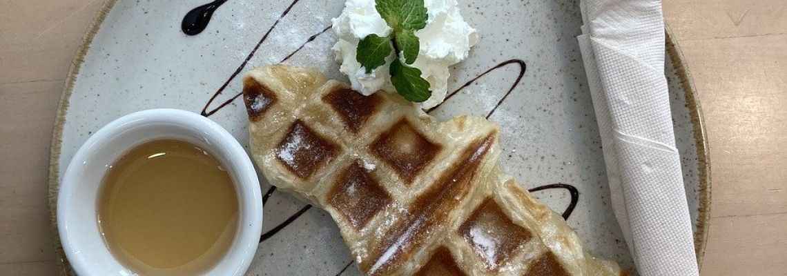 Een wafel met poedersuiker, slagroom en munt, met een kommetje siroop ernaast.