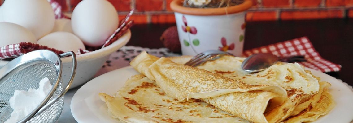 Dunne pannenkoeken op een bord, met eieren, bloem en een zeef ernaast.